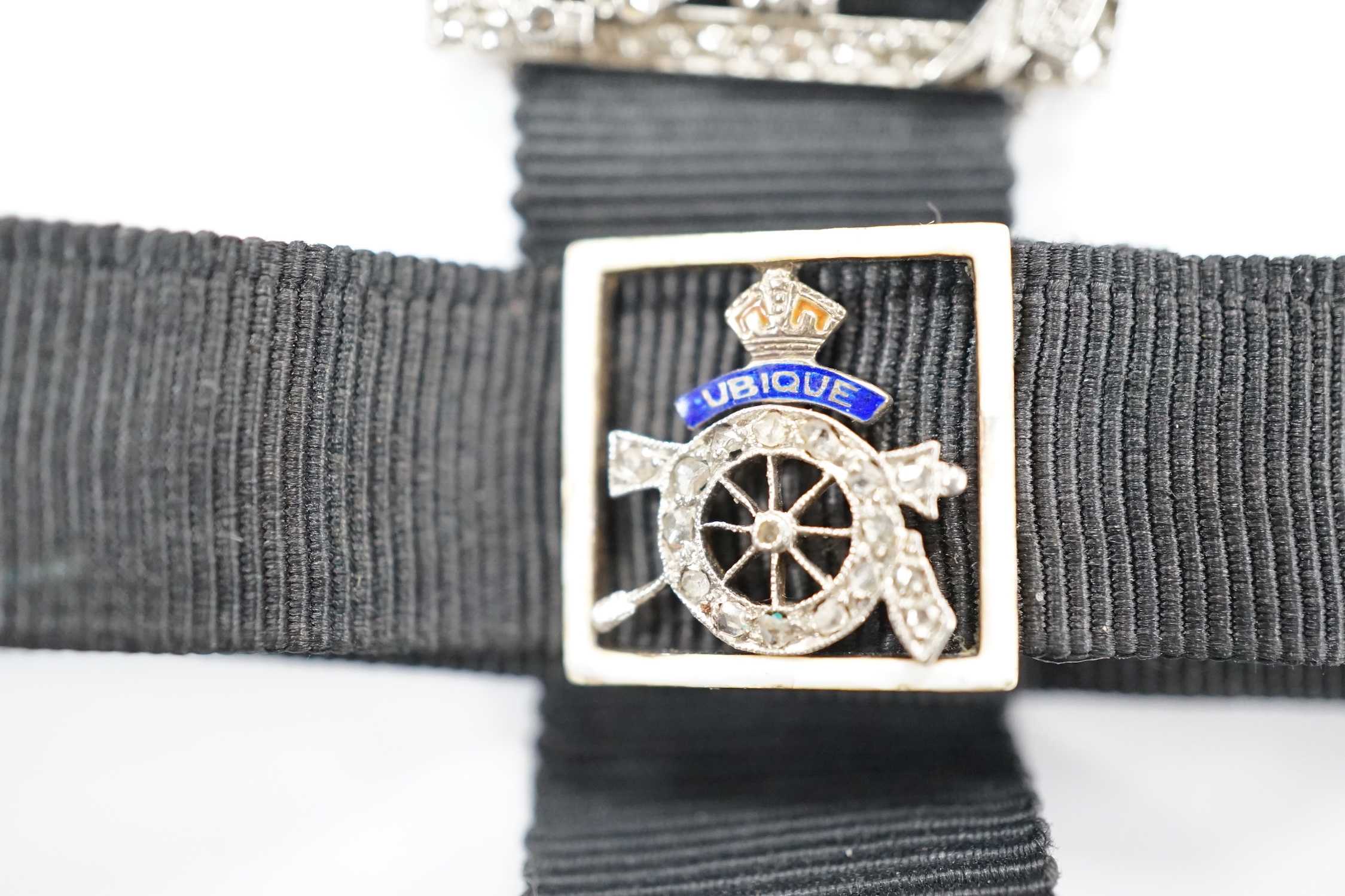 A black sash bracelet with diamond and enamel set Royal Fusiliers buckle, together with a similar sash with diamond set 'Charles' family crest, depicting a demi-rampant wolf, with motto.